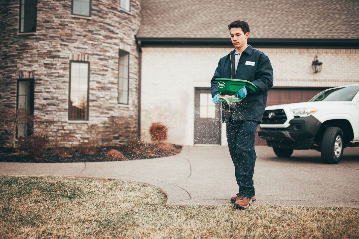 An Urbanex technician applying granular treatment to a front lawn, service truck in the driveway