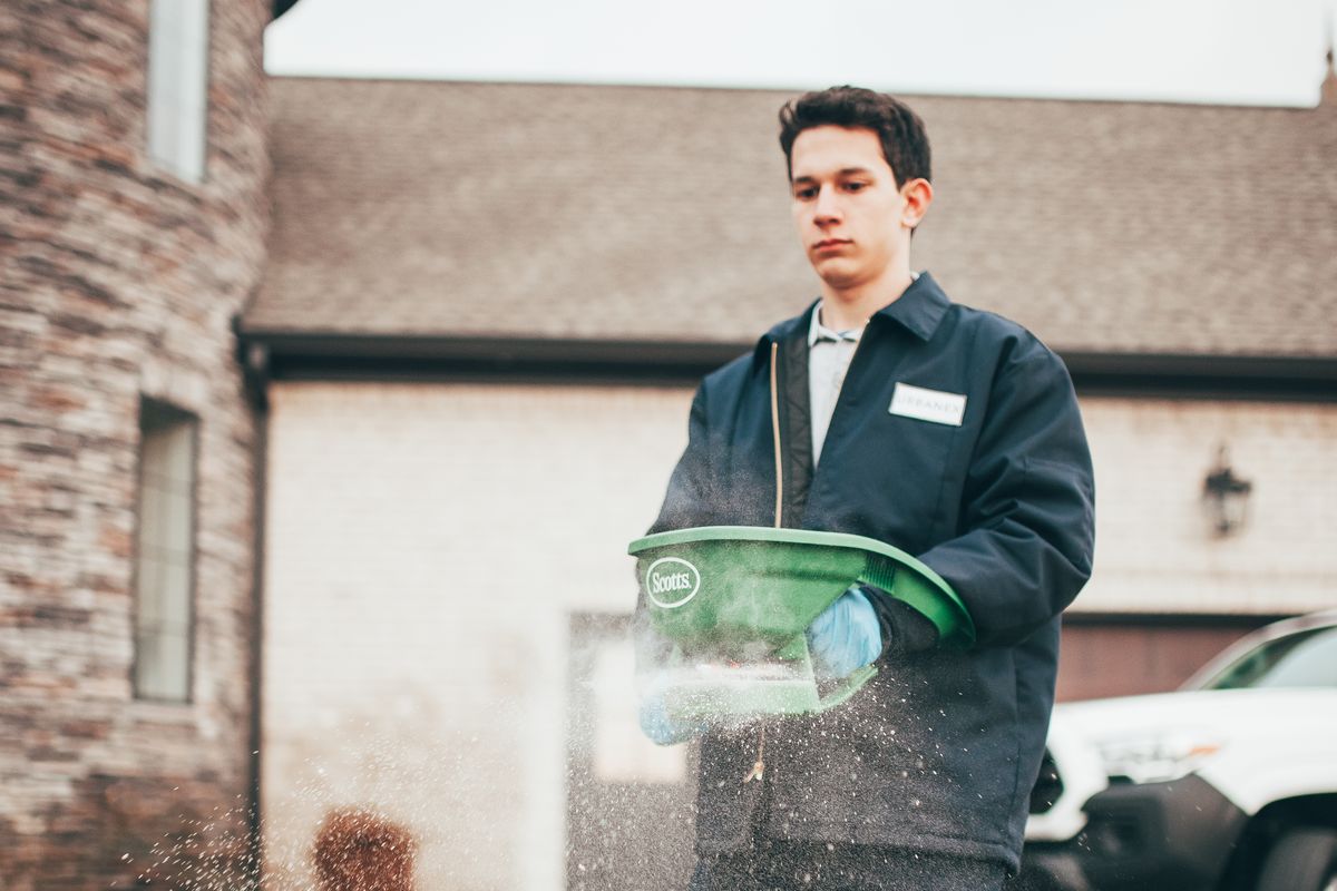 An Urbanex technician applying granular treatment to a lawn, service truck in the driveway behind