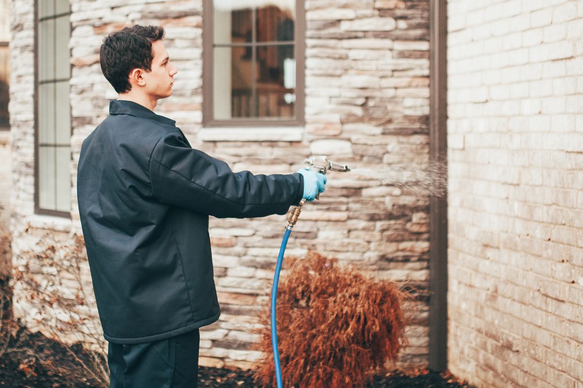 An Urbanex technician spraying a treatment along a home's stacked-stone exterior wall